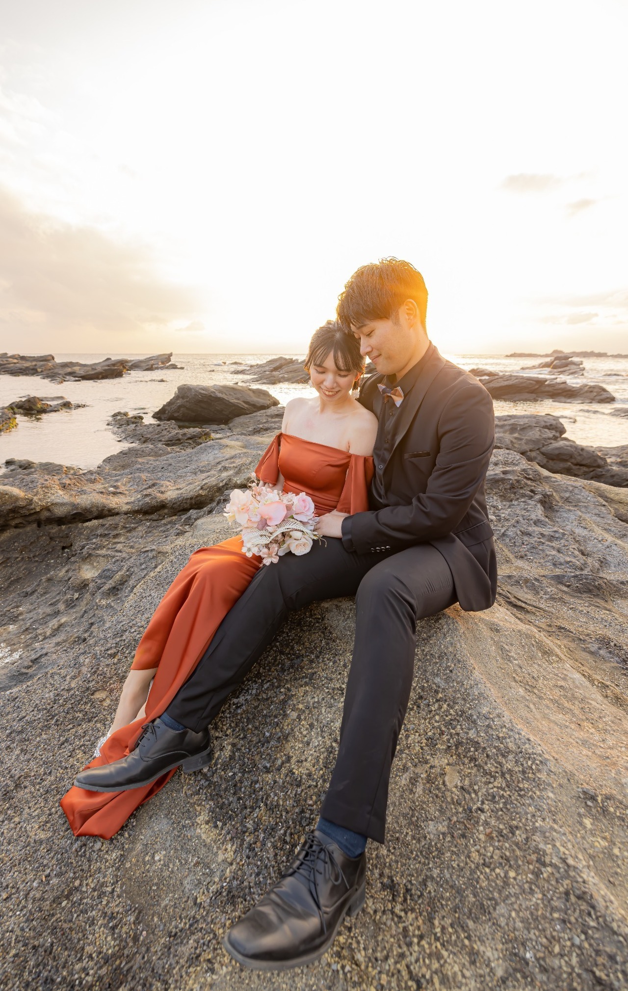 城ヶ島の夕陽を背に岩場で寄り添って座る新郎新婦の前撮り写真
