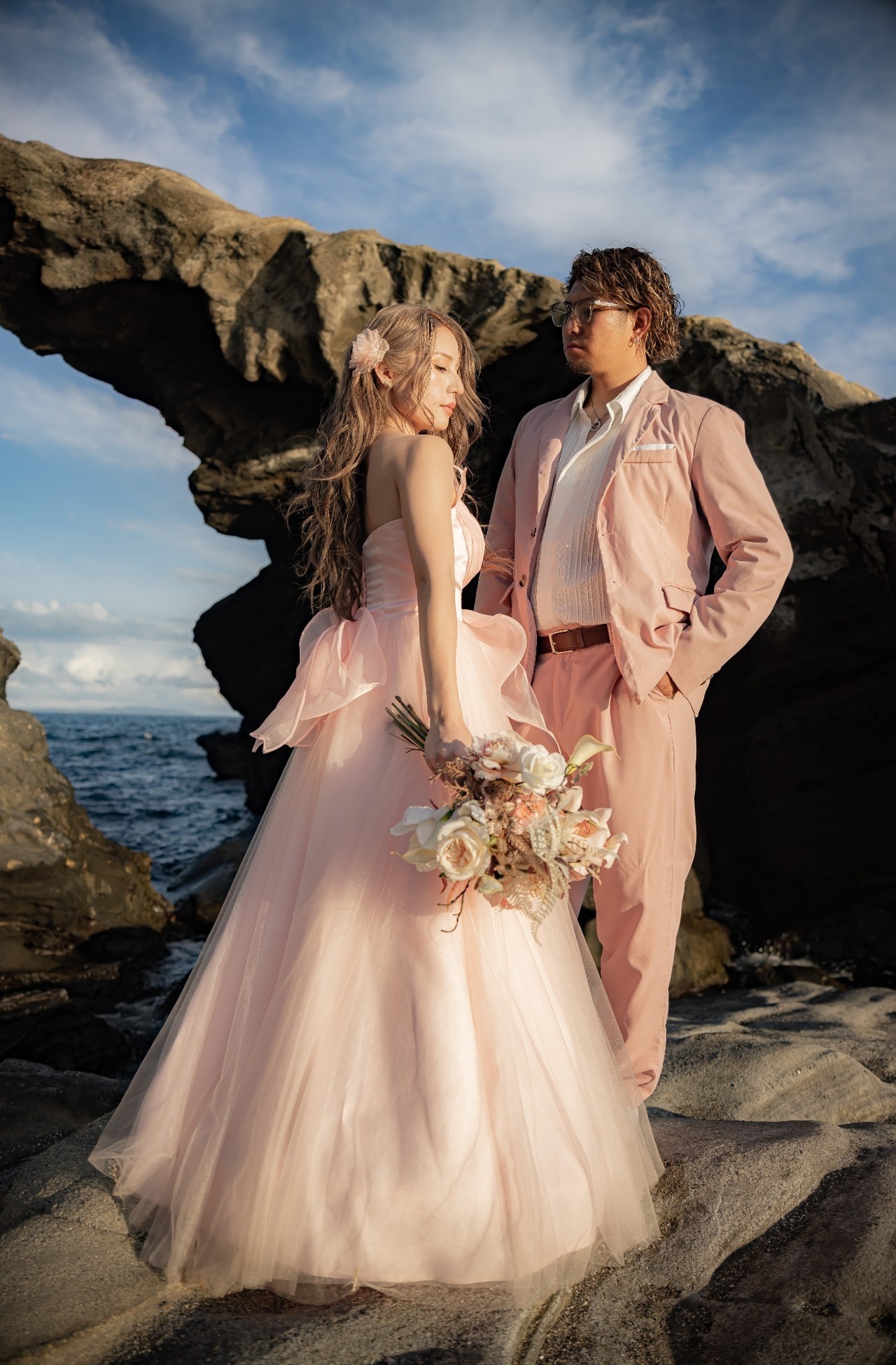 馬の背洞門を背景に、淡いピンクの衣装で立つ新郎新婦の前撮り写真