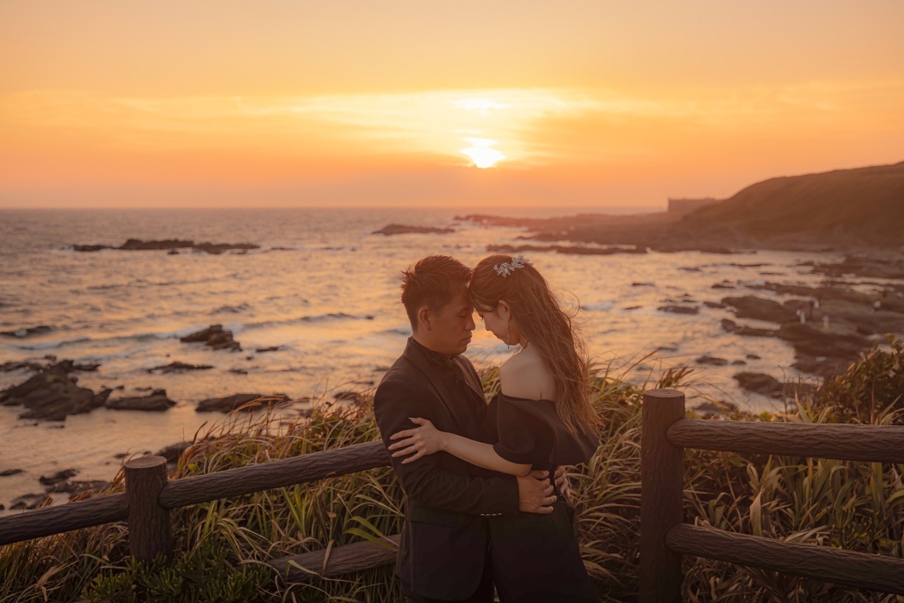 オレンジ色の夕陽と海を背景にブラック衣装で額を寄せ合う新郎新婦