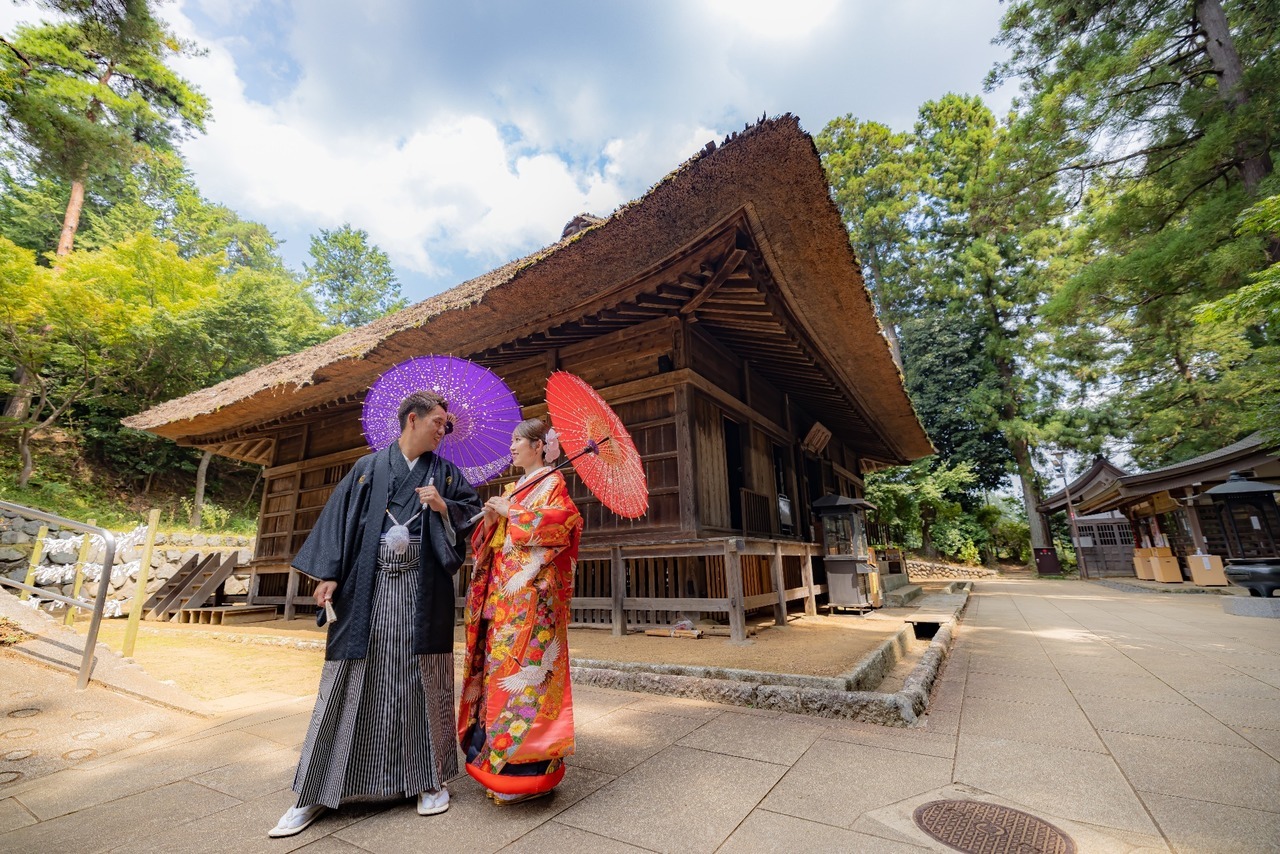 歴史を感じる木造建築の前で和傘を持ち見つめ合う色打掛と紋付袴姿の新郎新婦