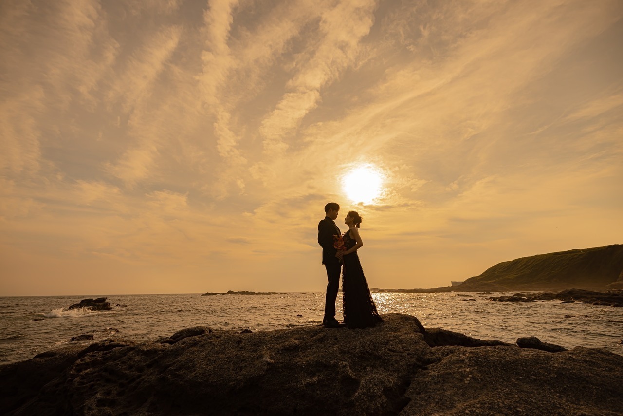 城ヶ島の夕日と海を背景に岩場で寄り添う新郎新婦の前撮りシルエット