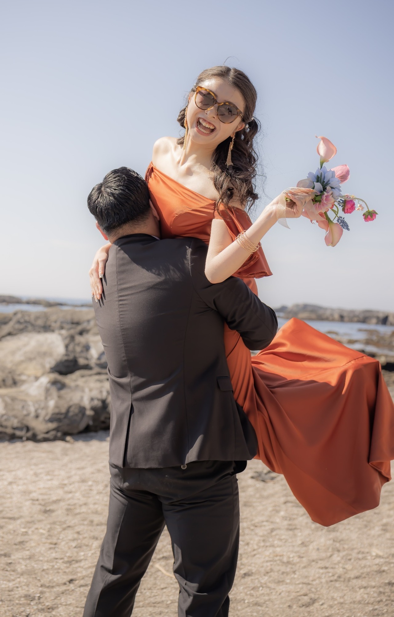城ヶ島の岩場で新郎に抱き上げられて笑うオレンジドレス姿の新婦